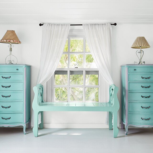 A cheery white-painted bedroom with a hint of blue-gray, a turquoise bench, and a window with white curtains flanked by turquoise-blue painted dressers with a lamp on each one.