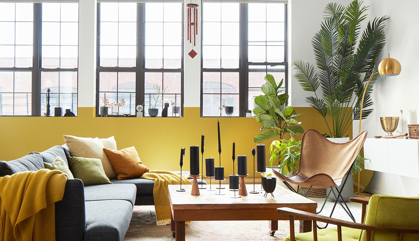 Un salon aéré, de style loft, présentant des murs et un plafond peints en blanc avec des tuyaux apparents, un mur d’accent blanc et jaune avec de hautes fenêtres, et un mobilier moderne gris, jaune et bois.