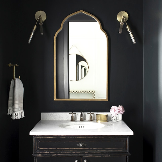 A sumptuous powder room with black-painted walls, brass accents, Moroccan tiled floors and white marble-topped vanity.