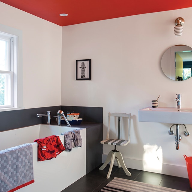 Salle de bains avec miroir rond et lavabo contemporain, murs noir et blanc accentués par un plafond rouge et une baignoire blanche.