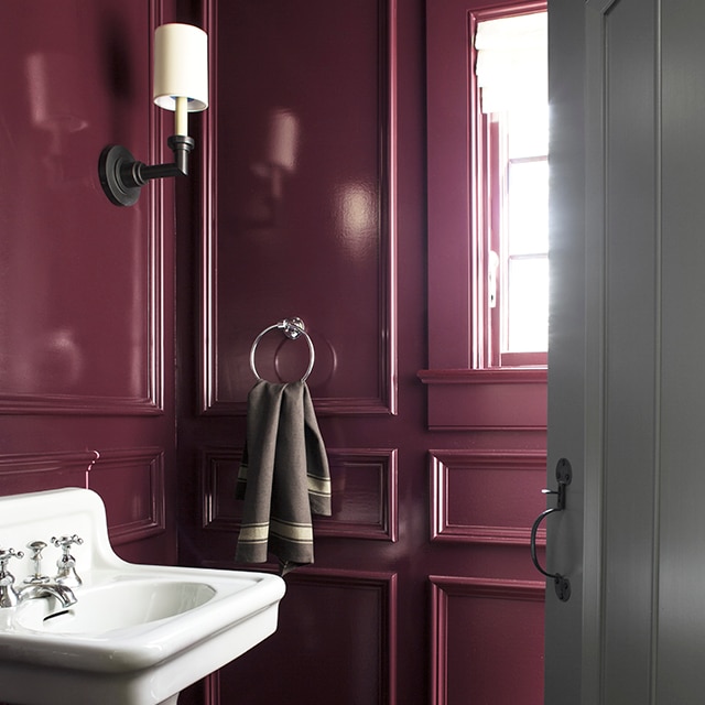 A bold powder room painted in a gloss dark plum hue with a freestanding white sink and gray-painted door.