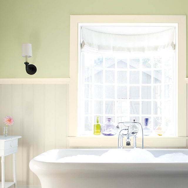 Magnifique salle de bains aux murs vert pâle avec lambris blanc cassé et baignoire remplie de bain moussant avec, au-dessus, une fenêtre panoramique aux boiseries blanches.