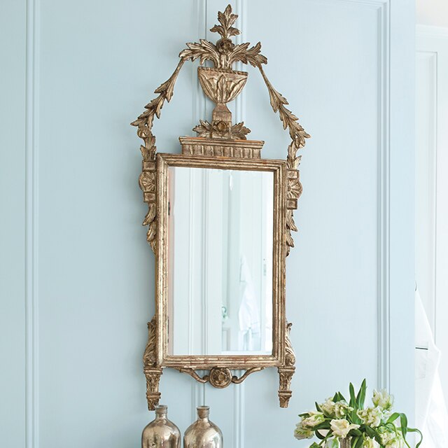 A close up on a vintage mirror against a light blue-painted bathroom, elegant polished sideboard holding silver bottles and a vase with white flowers. 