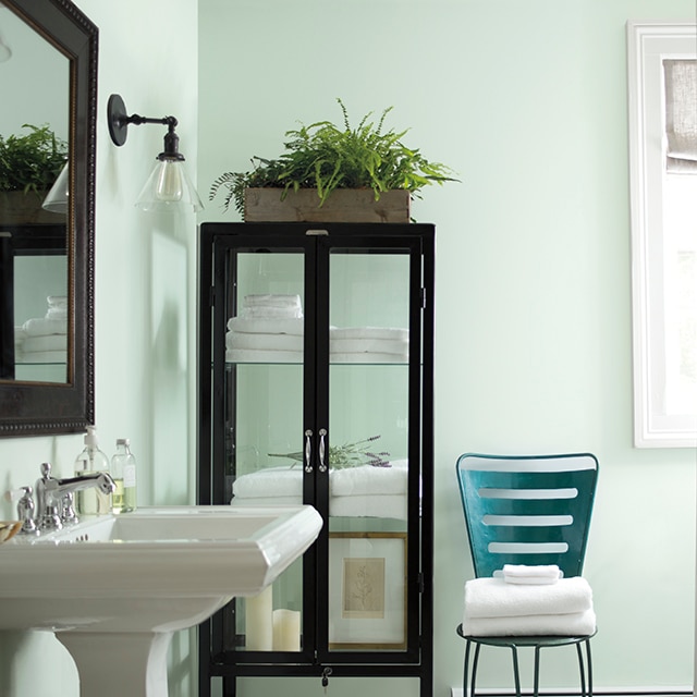 Salle de bains rafraîchissante vert pâle avec meuble d’apothicaire en verre, lavabo sur pied, chaise bleu sarcelle et miroir avec cadre en bois.