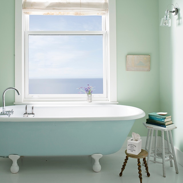 A serene light green bathroom with a light blue-painted clawfoot tub underneath a light-filled window with white trim.