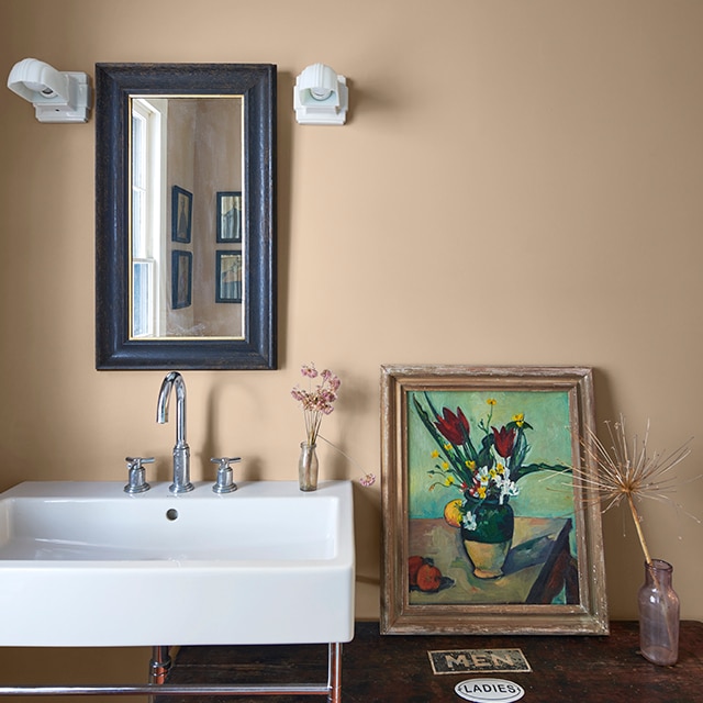 Un baño inspirado en la naturaleza con paredes pintadas de color bronceado, un espejo con marco negro, un lavabo blanco, una pintura de flores al óleo y dos pequeños jarrones con flores secas.