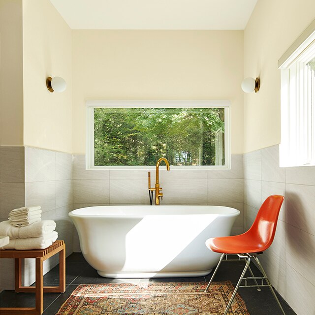 An off-white bathroom with a freestanding bathtub underneath a wide window, area rug, wood bench with towels, and red chair.
