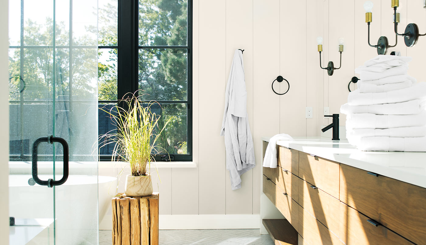 Matte bathroom with off-white painted shiplap walls, wooden vanity, over-sized windows with black painted trim, sisal bathmat, glass shower door and a planter on a small round wood table.