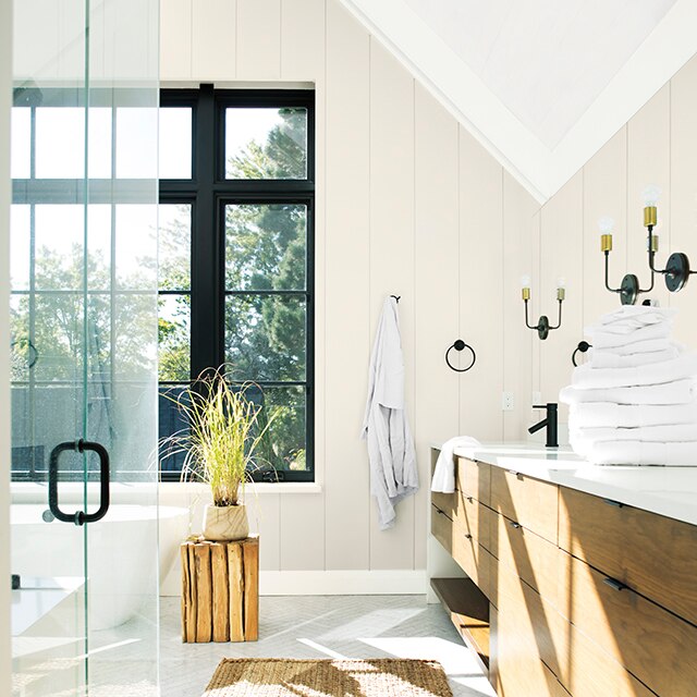 Matte bathroom with off-white painted shiplap walls, wooden vanity, over-sized windows with black painted trim, sisal bathmat, glass shower door and a planter on a small round wood table.