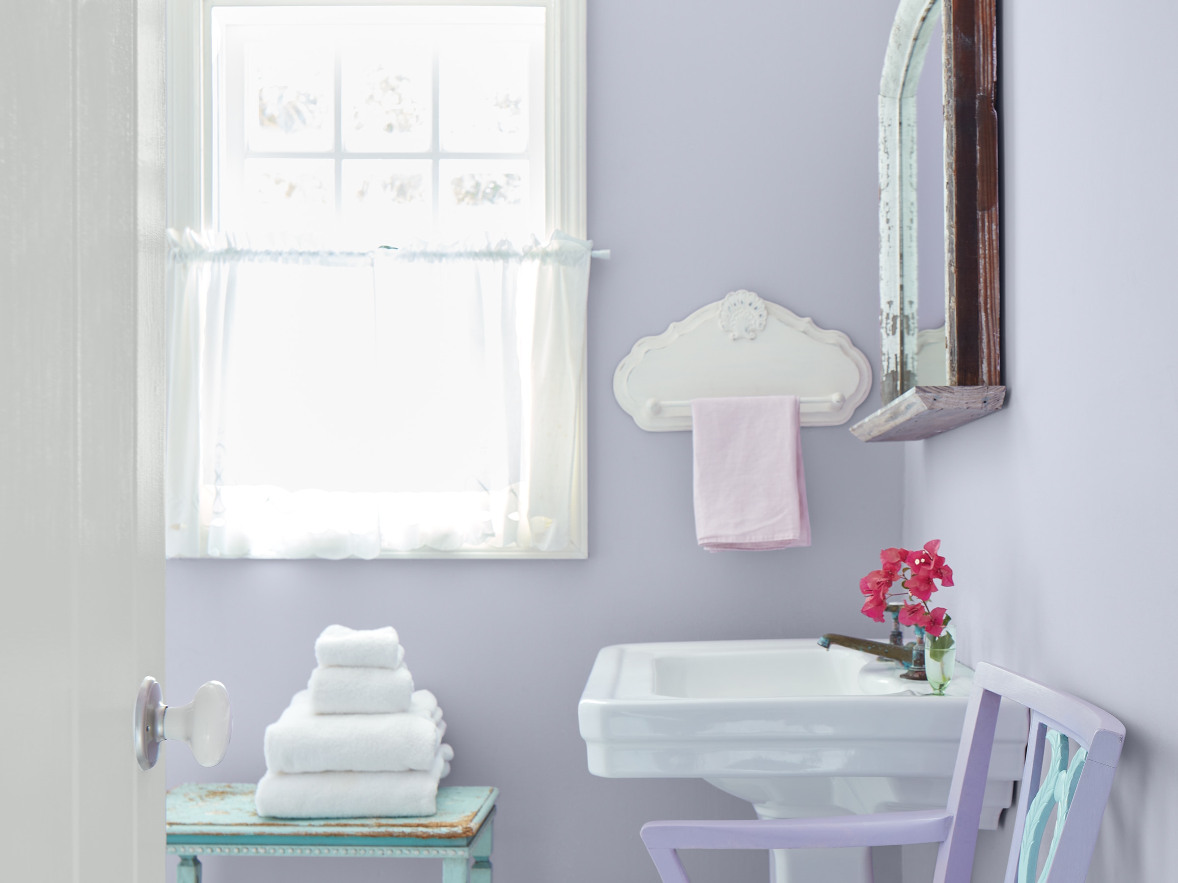 Un baño pequeño y soleado con paredes de color púrpura pastel, un lavabo blanco y una ventana con marco de blanco y cortinas blancas vaporosas.