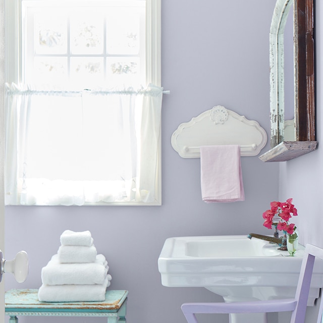 Un baño pequeño y soleado con paredes de color púrpura pastel, un lavabo blanco y una ventana con marco de blanco y cortinas blancas vaporosas.