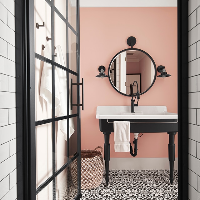 A subway tiled hallway and black-trimmed glass door open into a refreshing light pink-painted bathroom with black and white tiled floors and a wide farmhouse sink with a black base.