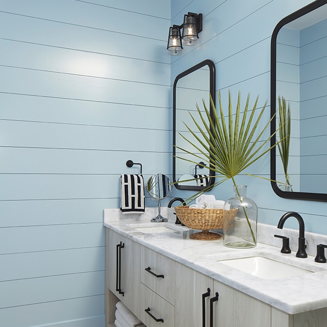 A bathroom with blue-painted shiplap walls, a white ceiling, a blonde wood double vanity with a marble top and black fixtures, and two mirrors.