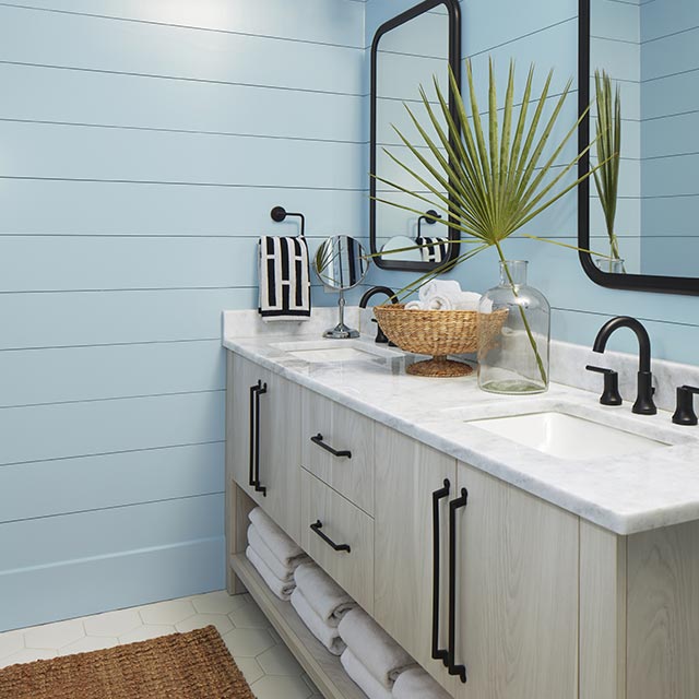Salle de bains aux murs en planches à feuillure bleues, plafond blanc, double meuble-lavabo en bois blanc avec comptoir en marbre, accessoires noirs et deux miroirs.
