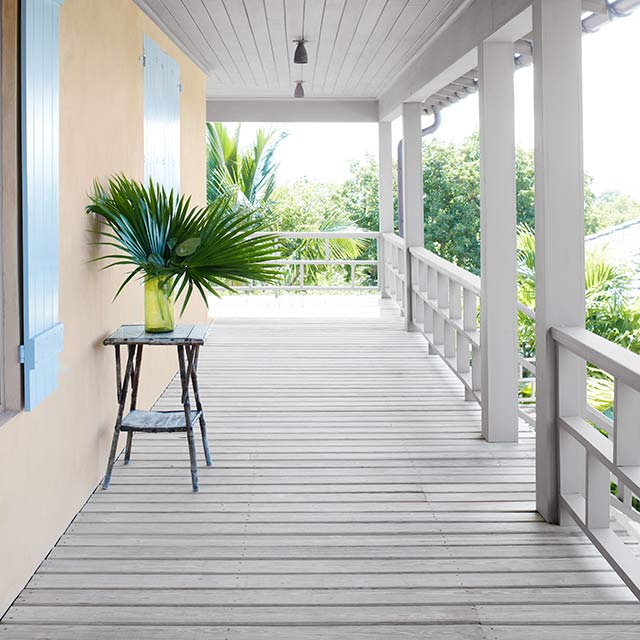 A cheery covered porch with a tan wall with blush undertones, blue shutters, gray stained ceiling, posts, railings and floor, palm leaves on a table, surrounded by greenery.