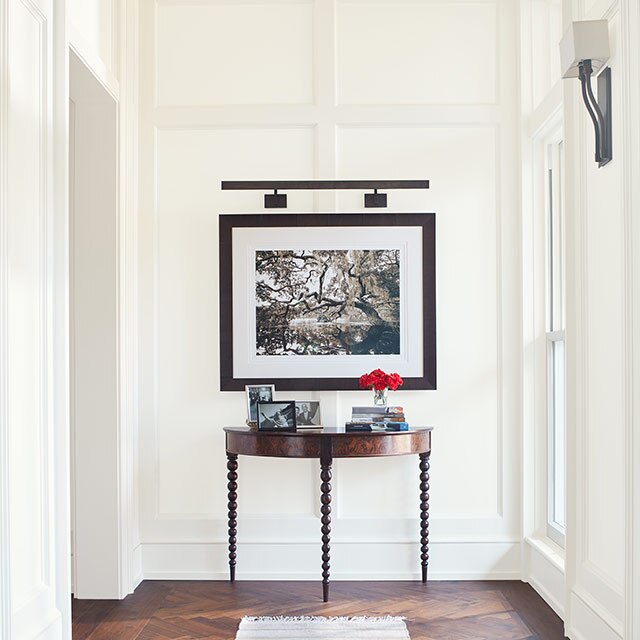 A bright, white-painted hallway with elegant board and batten walls, a piece of framed art, a table, tan striped rug, and dark wood floor.