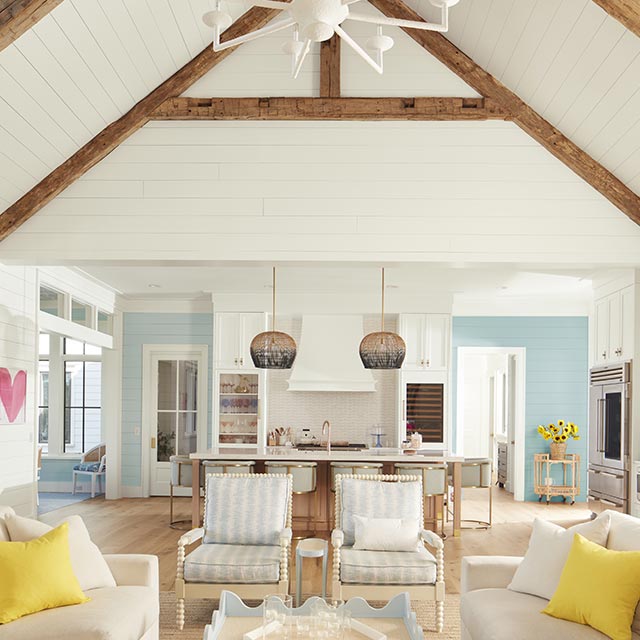 An airy, open white-painted living room and kitchen with shiplap walls and vaulted ceiling, wood beams, blue and white decor, and far back blue walls.