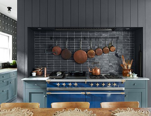A warm, inviting kitchen with blue-painted cabinets, a black shiplap wall and black backsplash, a white ceiling, blue stove, and wood table and chairs.