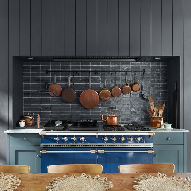 A warm, inviting kitchen with blue-painted cabinets, a black shiplap wall and black backsplash, a white ceiling, blue stove, and wood table and chairs.