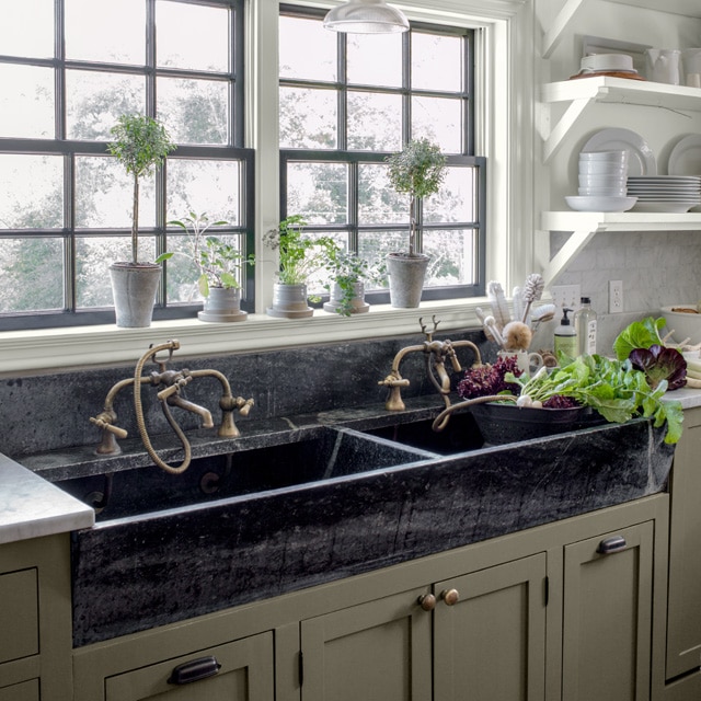 A double black kitchen sink beneath paned windows, and lower deep sage green-painted cabinetry and an off-white wall, open shelving and ceiling.