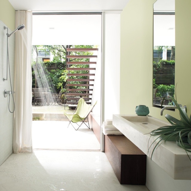 Chambre contemporaine vert pâle aux lignes épurées avec plafond blanc, jet d’eau de la douche grand ouvert et grande porte ouverte sur un espace extérieur illuminé par le soleil.