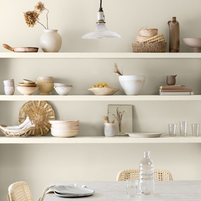 White-painted shelves holding baskets, bowls, ceramics, glasses and other off-white décor against a white-painted wall, and a table with two chairs. 