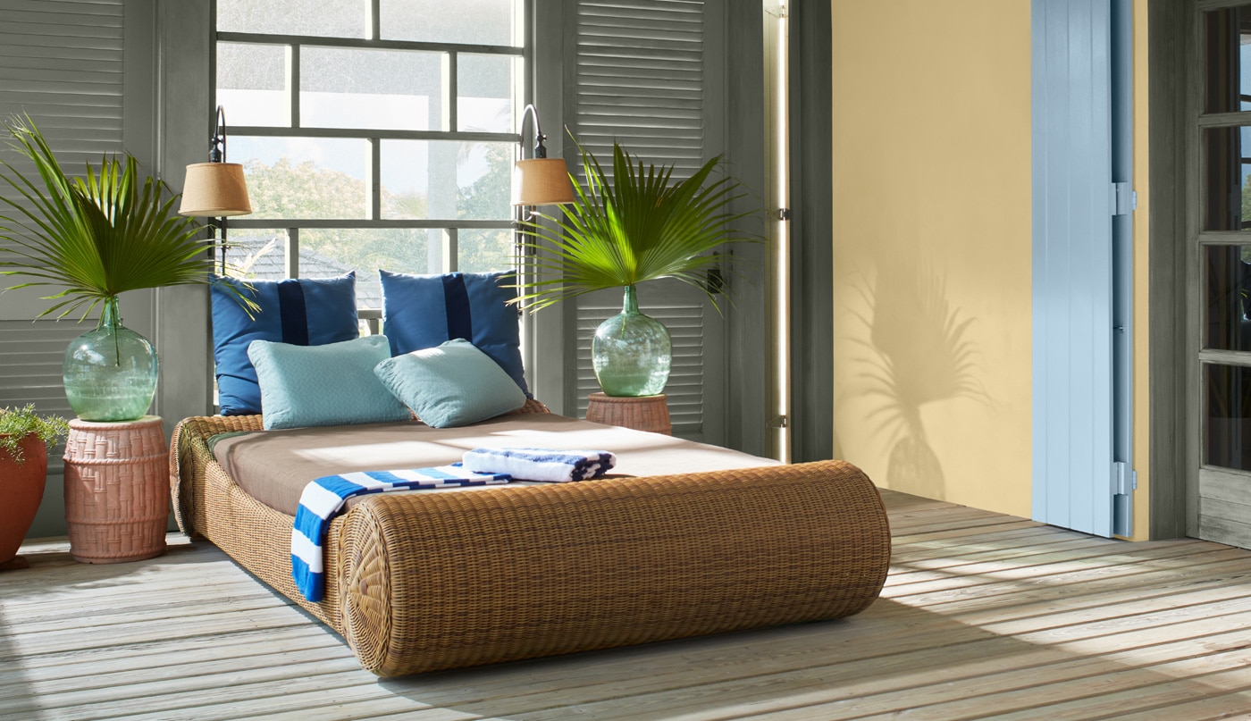 A beautiful bedroom with a yellow-painted accent wall, earthy green trim, doors and window, a light blue entrance door shutter, a rattan bedframe and palmettos in vases flanking both sides of bed.