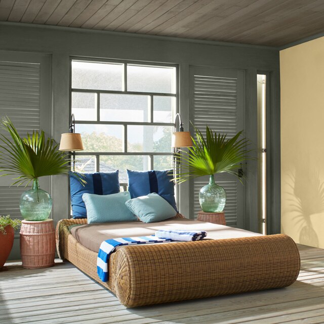 A beautiful bedroom with a yellow-painted accent wall, earthy green trim, doors and window, a light blue entrance door shutter, a rattan bedframe and palmettos in vases flanking both sides of bed.