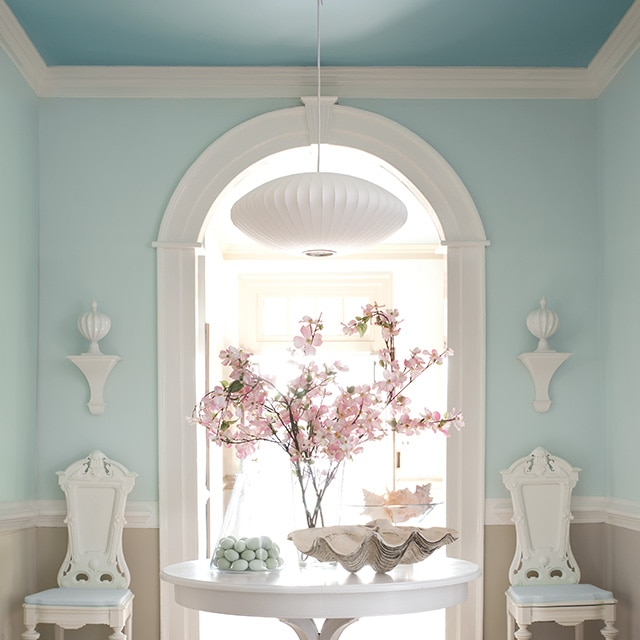 A spacious hallway with light blue painted upper walls, neutral lower walls, and a blue ceiling features a white table and chairs, and a white painted arch and trim throughout.  