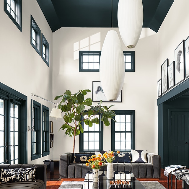 A sunlit living room with two-story white painted walls; a deep green vaulted ceiling, trim, and doors; large oval pendant lights; and neutral modern decor.