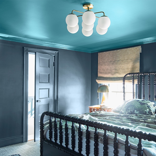 This cozy bedroom features a teal painted ceiling and crown molding; dark gray walls, trim, and door; and a spindle bed.