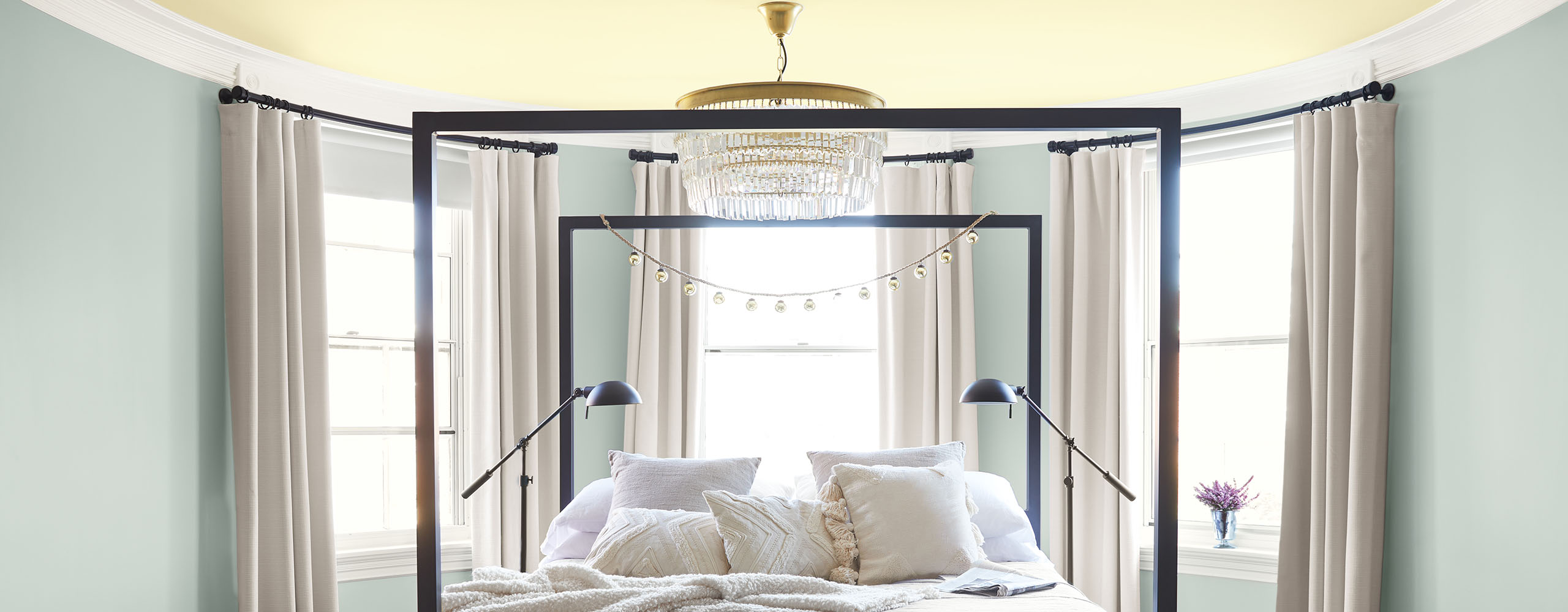 Rounded bedroom walls painted a soft blue-gray, a pale yellow ceiling, white trim, and three windows surround a comfy black four-poster bed.