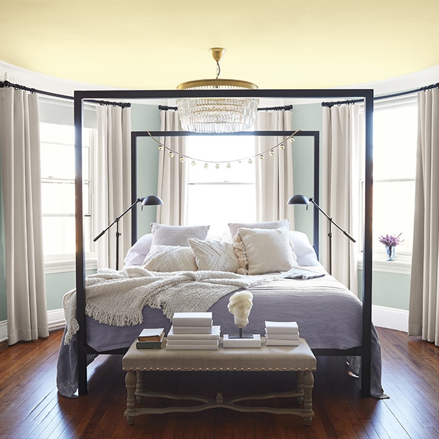 Rounded bedroom walls painted a soft blue-gray, a pale yellow ceiling, white trim, and three windows surround a comfy black four-poster bed.