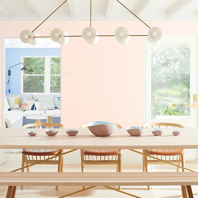 An airy, open dining room with a light pink painted wall, a white beamed ceiling, white french doors, and modern wood dining furniture, looking into a blue-painted living room.