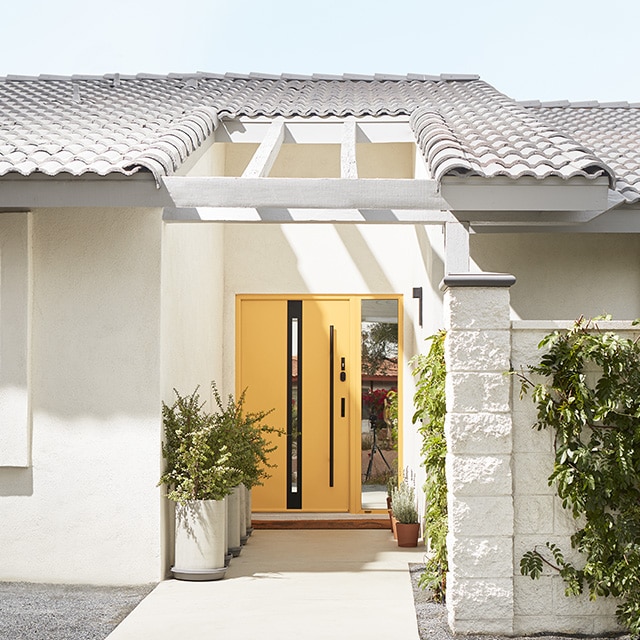 A sunny home exterior painted in off-white with a yellow front door, cacti and other desert landscaping.