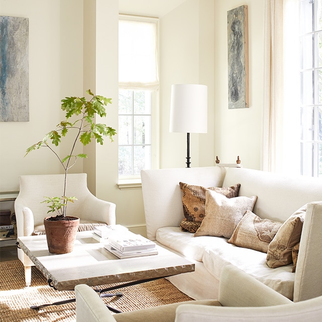 An inviting, sunlit, white-painted living room with a comfy white sofa and two chairs, and a stone top table with a potted plant.