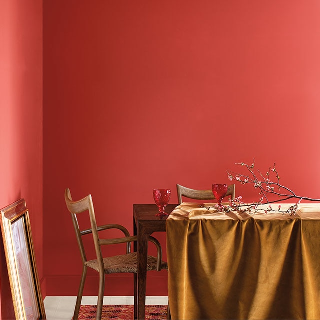 A dining room corner with bold coral painted walls with a hint of pink, wood chairs, a gold cloth draped over a table, and a picture leaning against one wall. 