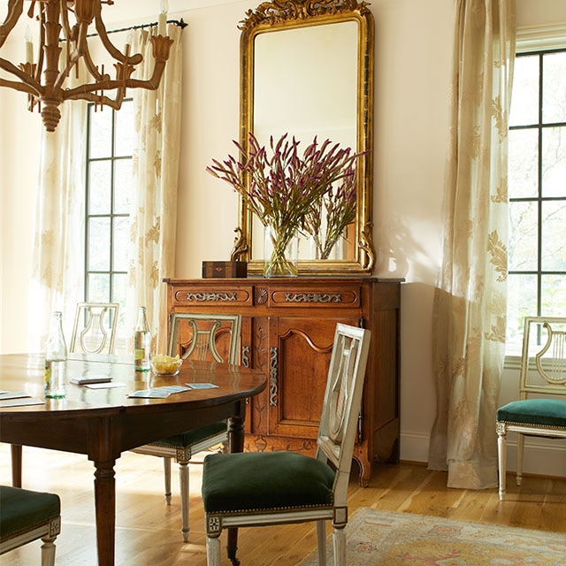 An elegant style dining room with a warm, off-white painted wall and ceiling, mixed wood furniture, a gold mirror and chandelier, beige drapes, and gold area rug.