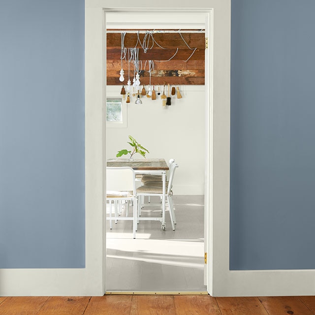 Un couloir aux murs bleus avec moulures et plafond blancs donne sur une salle à manger blanche lumineuse au plafond avec poutres en bois, des meubles blancs de style épuré et un dessus de table en bois. 