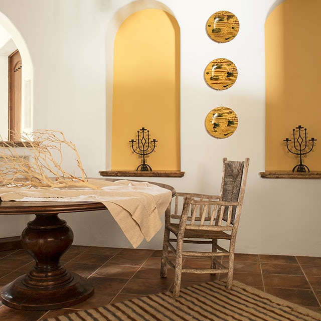 An airy, white-painted dining room with two yellow-painted alcoves, yellow decorative plates, dark wood ceiling beams, a rustic metal chandelier, and a round wood table and chair.