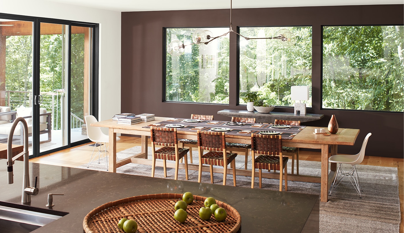 An open, modern dining room with a brown-painted accent wall and three large windows, a white ceiling and side wall with sliding glass doors, and brown granite kitchen island in the forefront. 