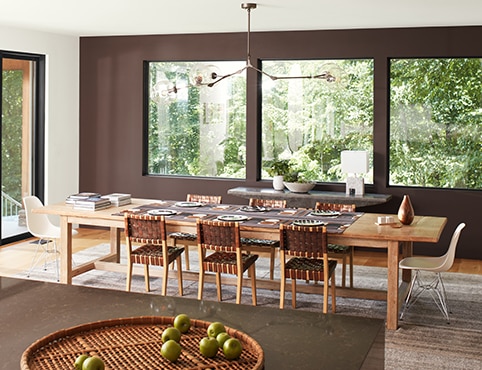 An open, modern dining room with a brown-painted accent wall and three large windows, a white ceiling and side wall with sliding glass doors, and brown granite kitchen island in the forefront.