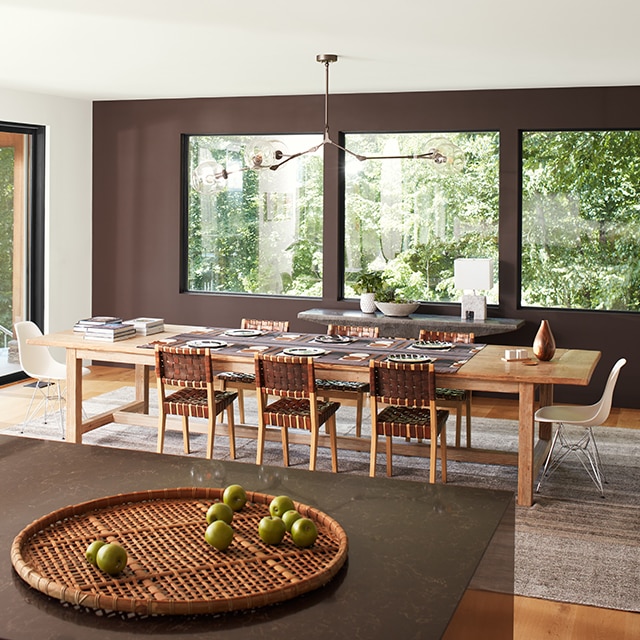 An open, modern dining room with a brown-painted accent wall and three large windows, a white ceiling and side wall with sliding glass doors, and brown granite kitchen island in the forefront. 