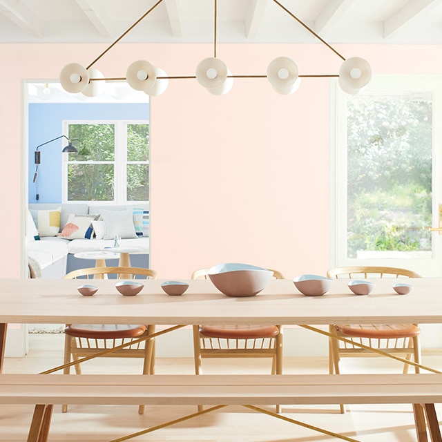 A light pink painted dining room with a white beamed ceiling, white glass doors, a wooden table, chairs, and bench, and a blue painted room in the background. A light pink painted dining room with a white beamed ceiling, white glass doors, a wooden table, chairs, and bench, and a blue painted room in the background.