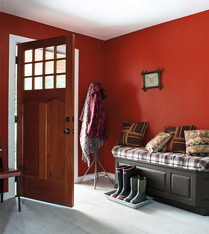 A deep red entryway does double duty as  mud room with several pairs of boots lined up next to an upholstered bench.