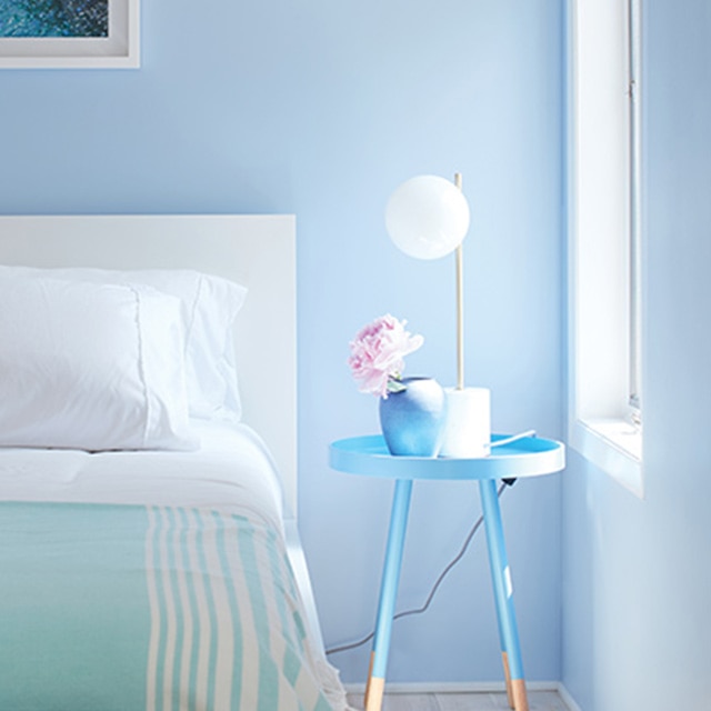 Chambre à coucher avec murs bleus et moulures blanches, couverture vert écume de mer à rayures blanches, tête de lit blanche et table bleue.