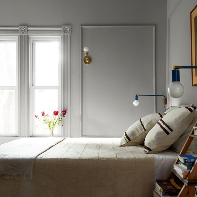 A bedroom with greige-painted walls, picture frame wall molding, and decorative window trim, a white ceiling and off-white décor and bedding.