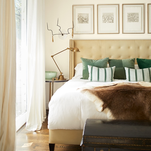 An off-white-painted bedroom with white and green bedding and pillows, a tan upholstered headboard and off-white drapes. 