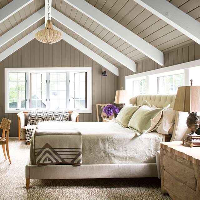 Chambre principale avec murs et plafond en voûte gris, moulures et poutres en bois blanches avec accessoires aux teintes neutres.