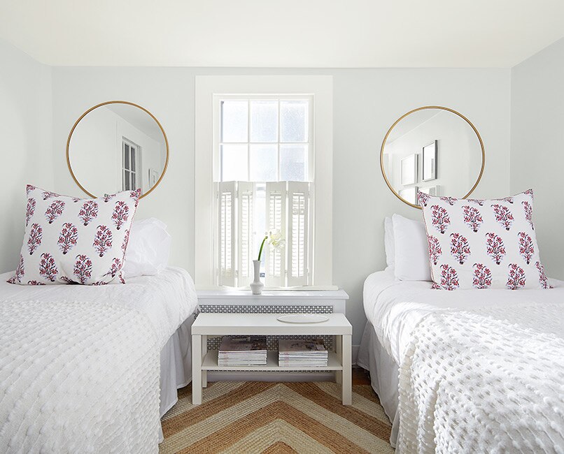 A white-painted bedroom with two circular wall mirrors, twin beds with white bedding, a white shuttered window and striped jute rug.